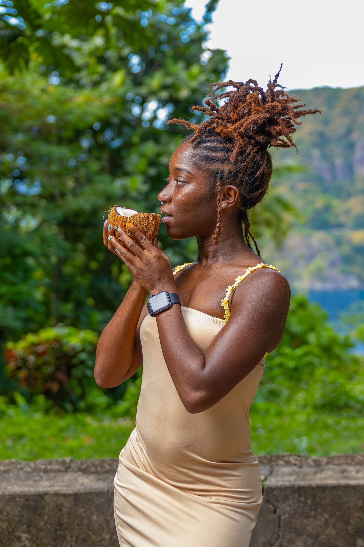 Woman in beige dress drinks from coconut while standing in tropical setting with ocean view.