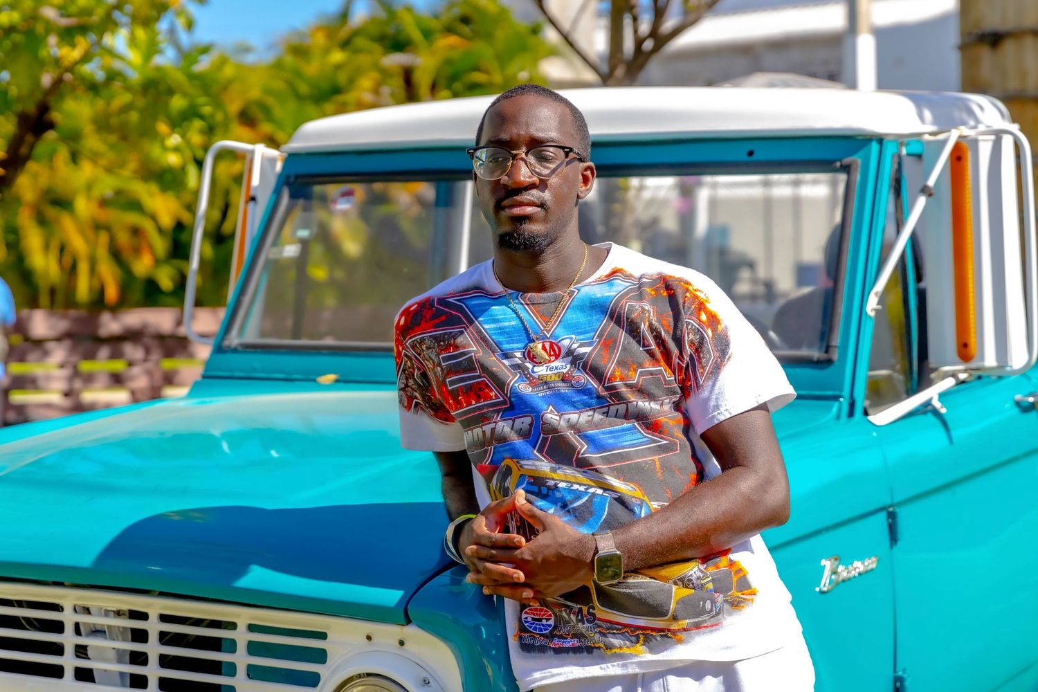 Someone wearing a colorful patterned shirt stands smiling in front of a vintage turquoise pickup truck.