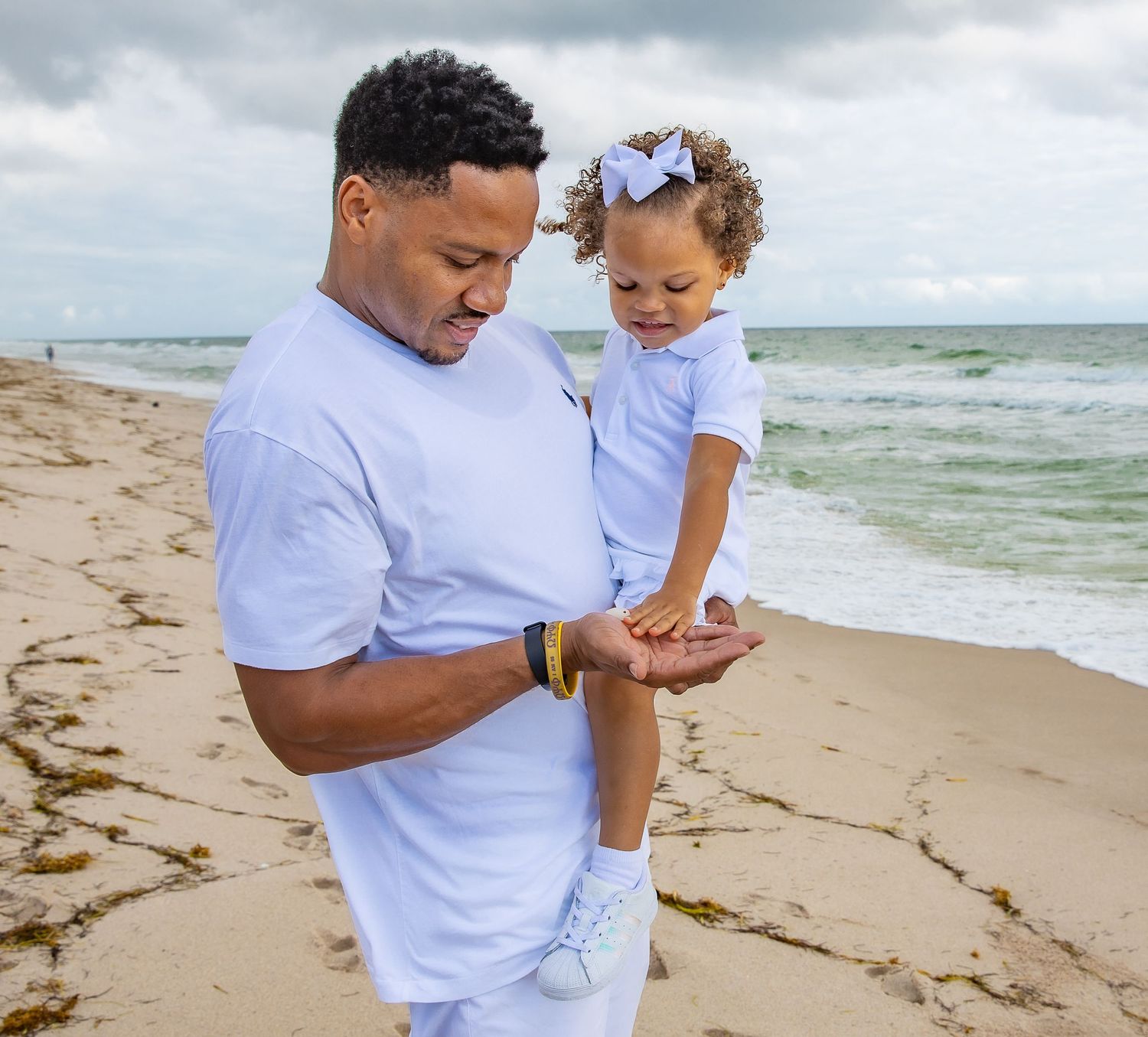 A toddler in a purple shirt and bow explores something in their parent's palm on a sandy beach by the ocean.