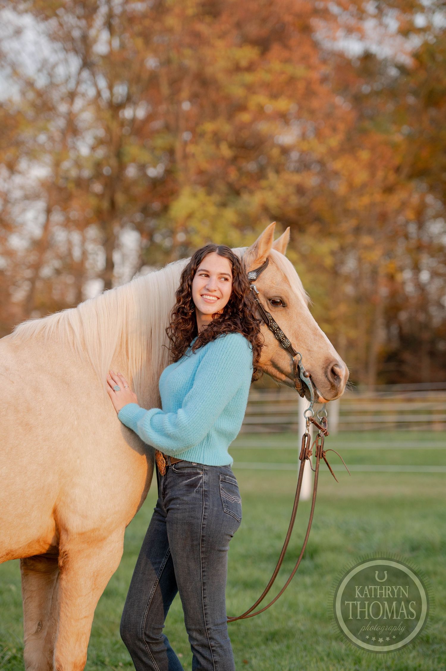 Someone in a blue sweater stands beside a cream-colored horse during fall season.
