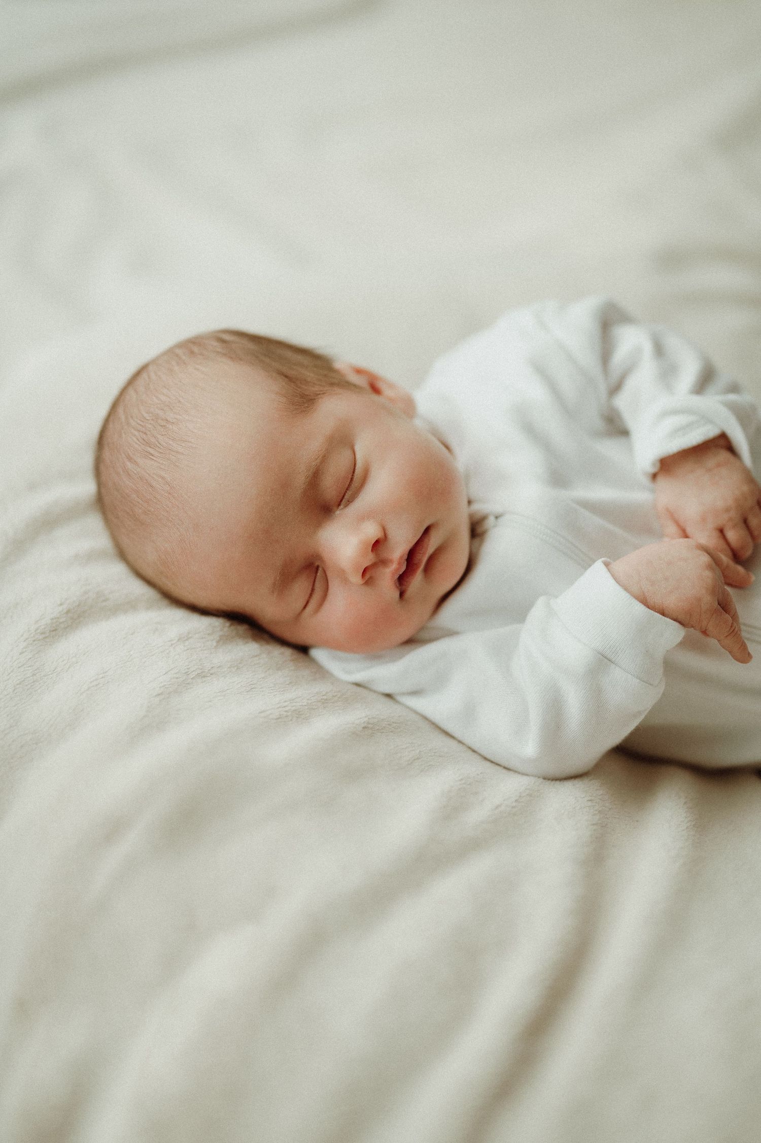 A peaceful sleeping newborn baby wearing a white outfit rests on light colored bedding.
