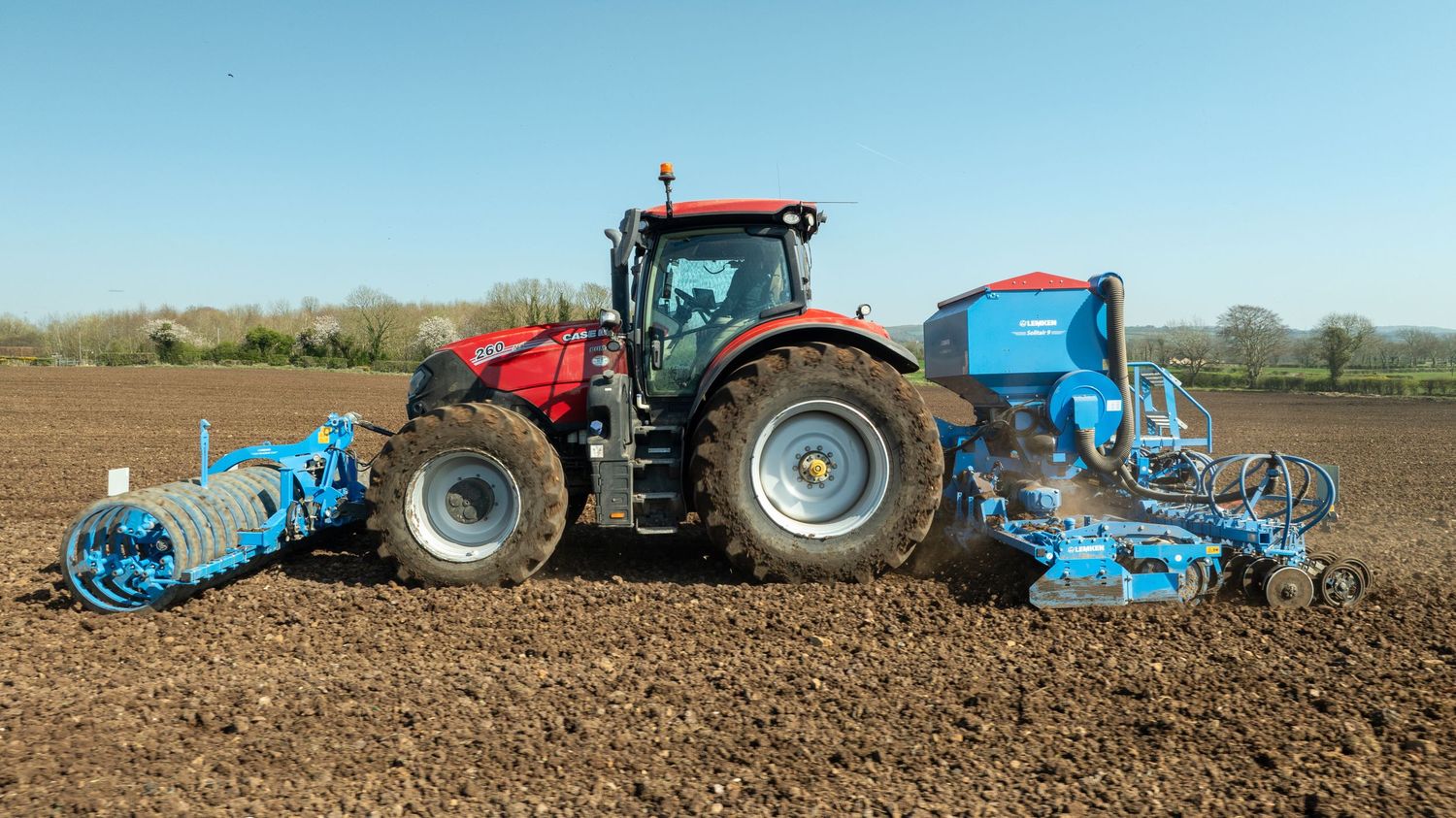 A red Case tractor with blue farming implements tills freshly plowed soil in a rural agricultural field.