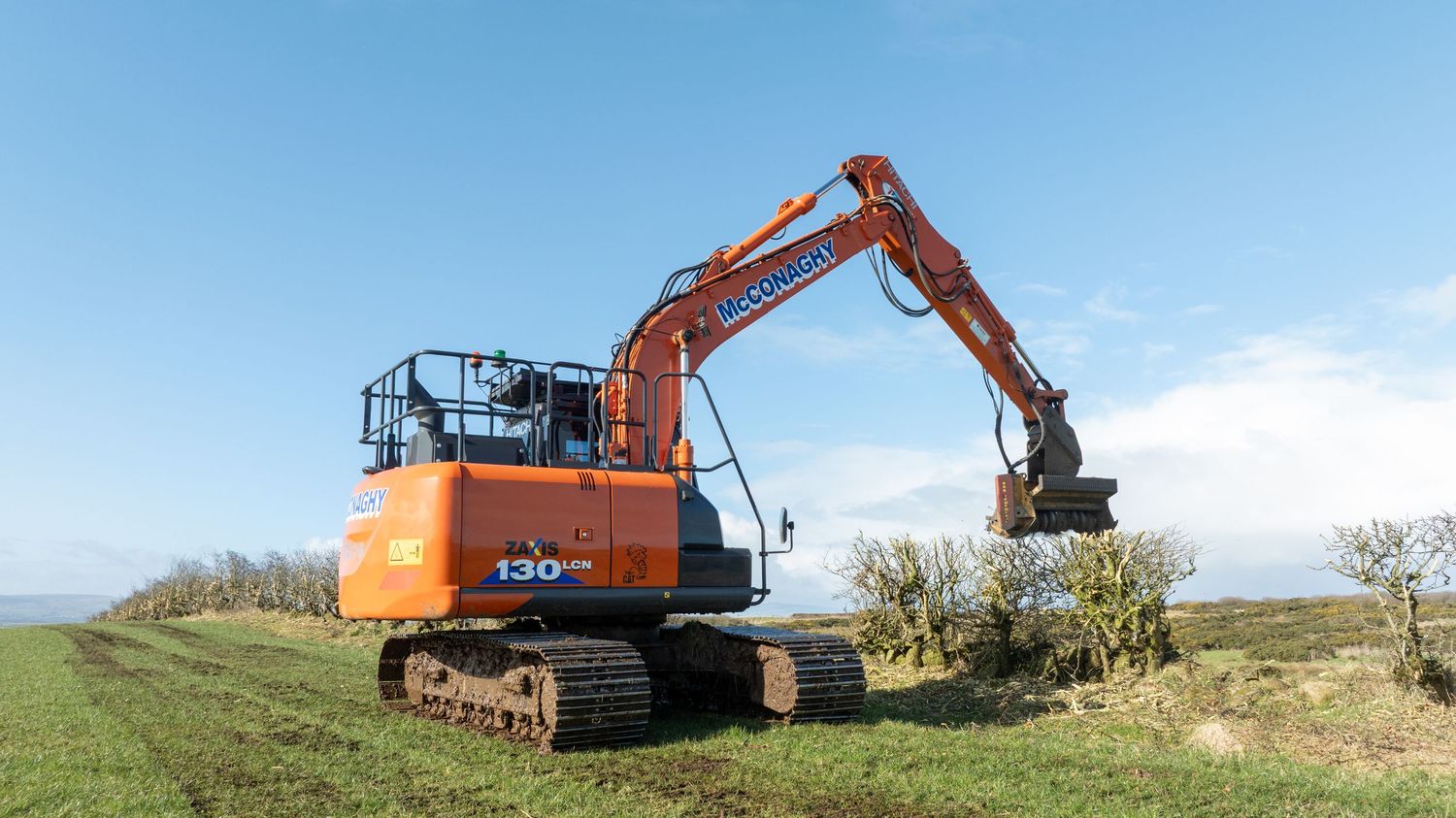 Orange excavator with mechanical arm working in rural field clearing vegetation against blue sky.