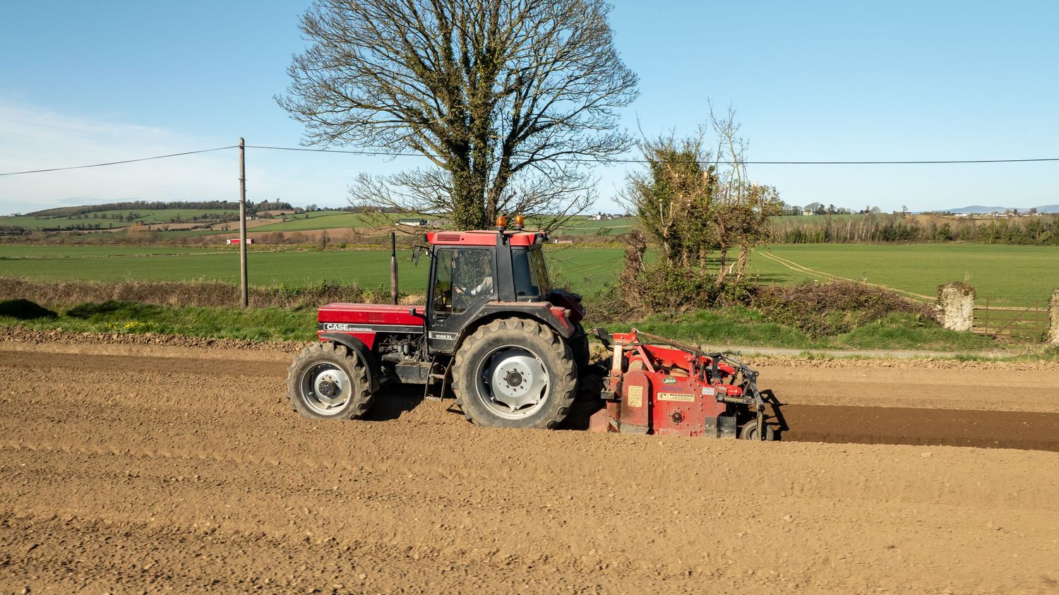 A red tractor with cultivation equipment works through a freshly plowed field on a sunny spring day.