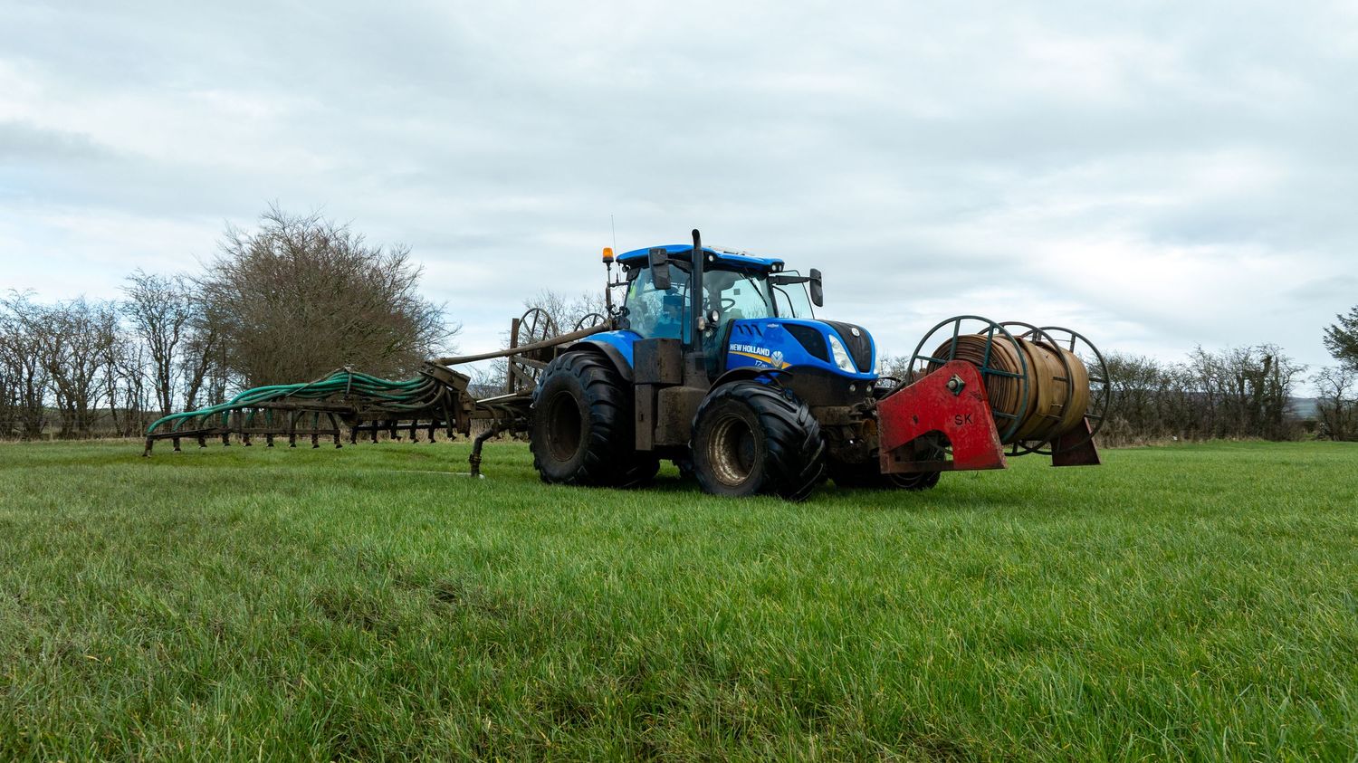 A blue New Holland tractor with red farming equipment attached works in a lush green field under cloudy skies.