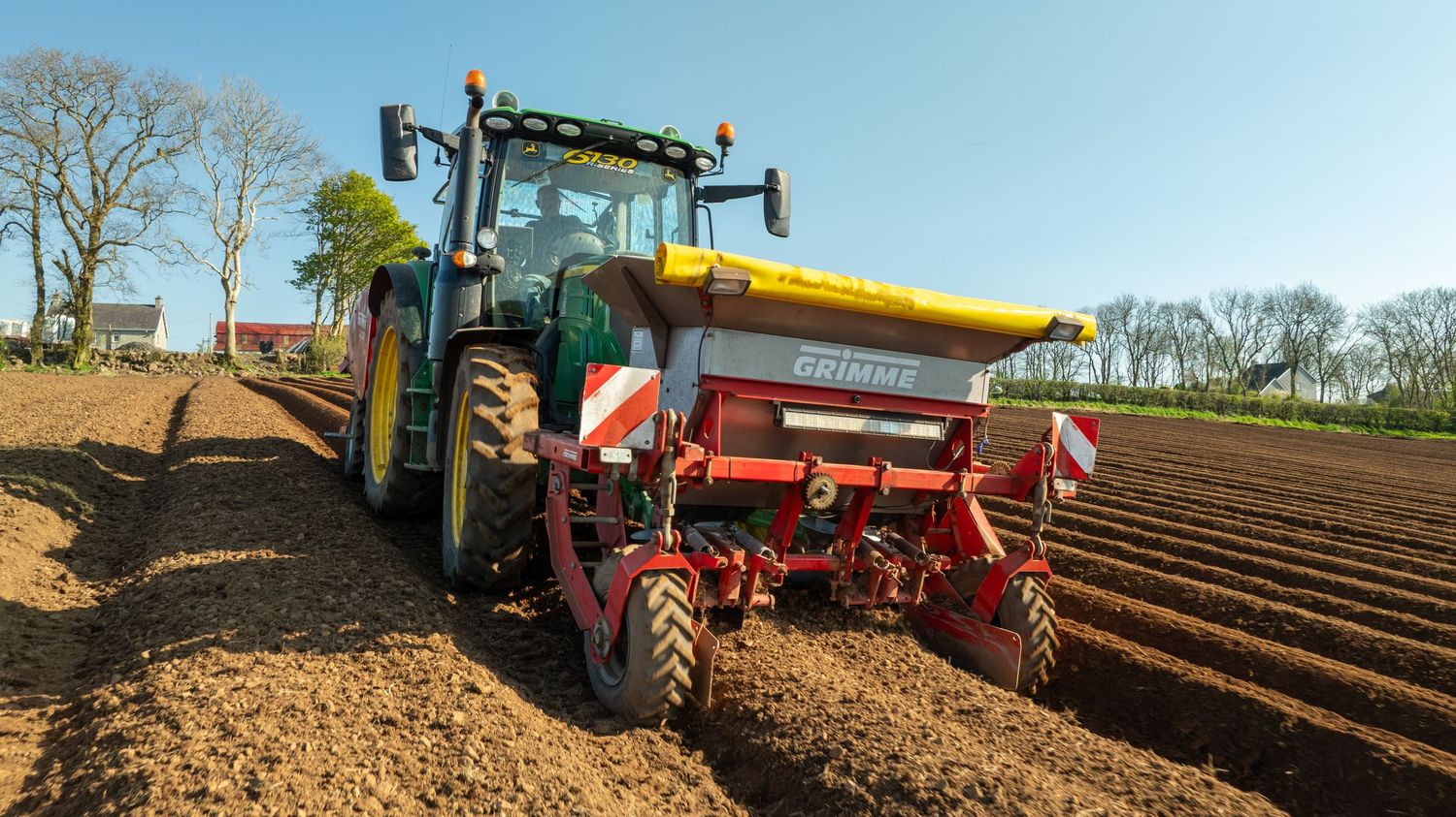 Green tractor pulling red potato planting equipment across freshly tilled soil in agricultural field during spring planting.