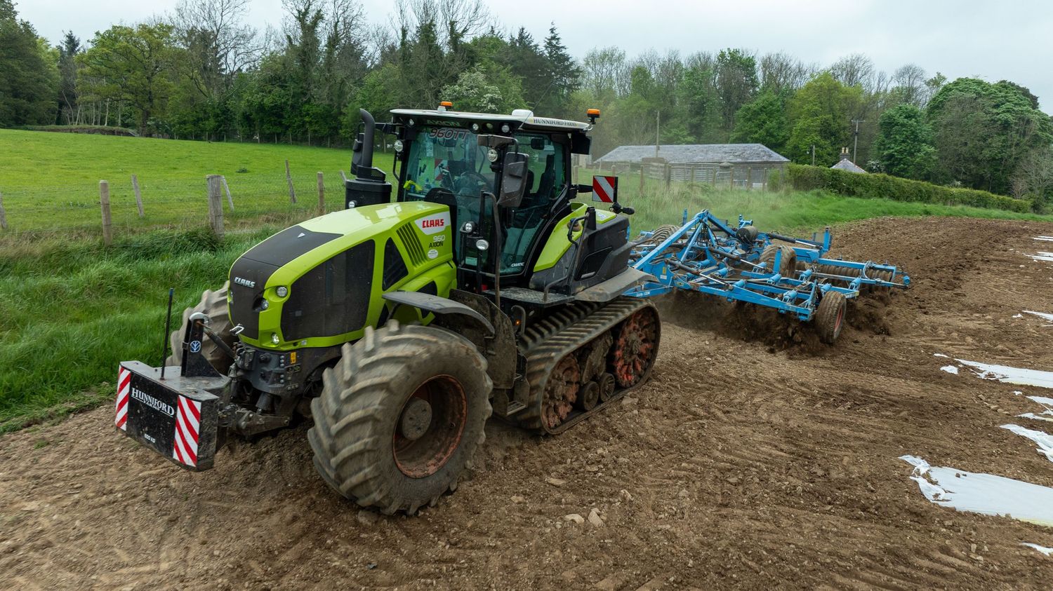 Large green tracked Claas tractor plowing a muddy field with attached farming equipment on a cloudy day.