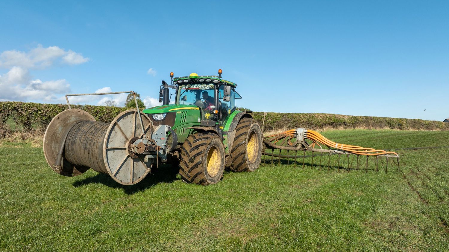Large green tractor with cable reel attachment and agricultural equipment on grassy farmland under blue sky.