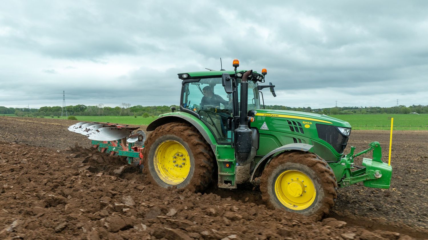 Green John Deere tractor pulling agricultural equipment across freshly plowed farmland under overcast skies.