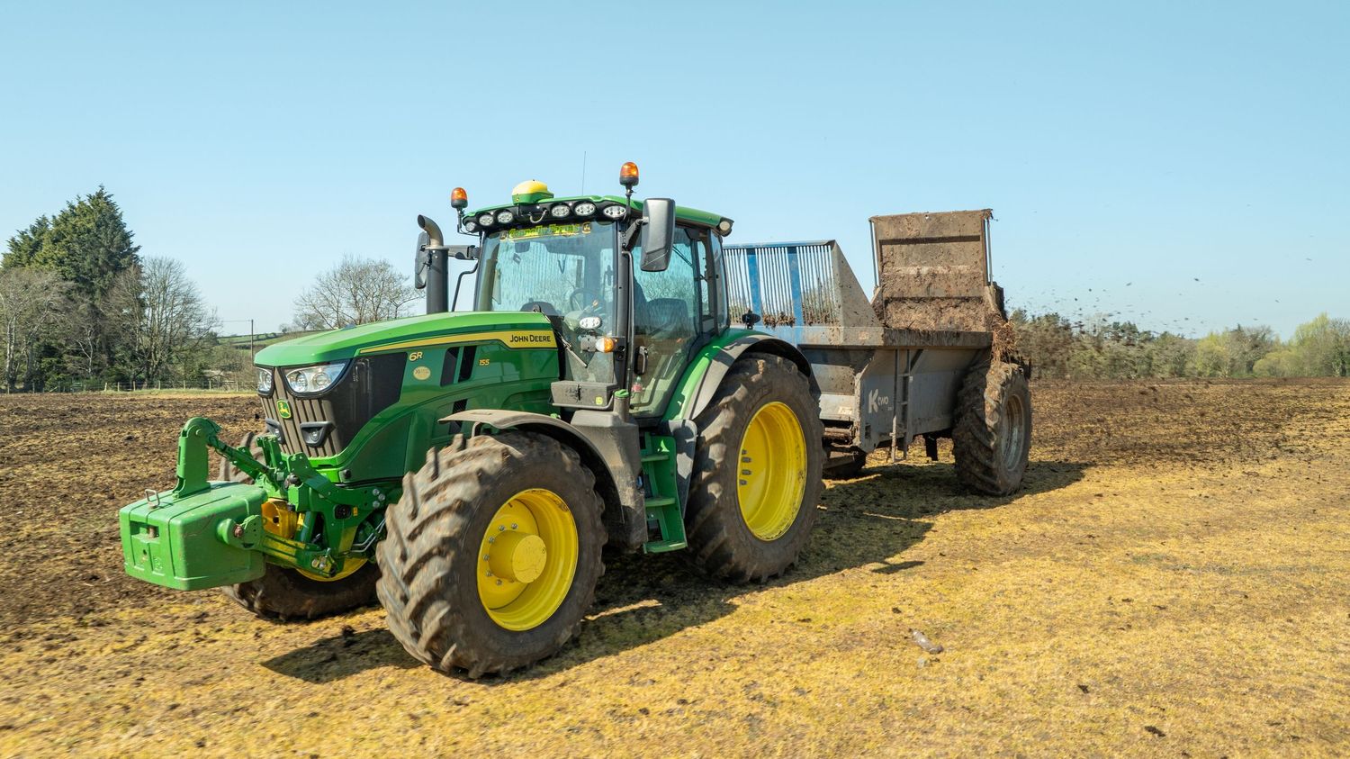 John Deere green tractor with attached brown trailer working in dusty agricultural field on sunny day.