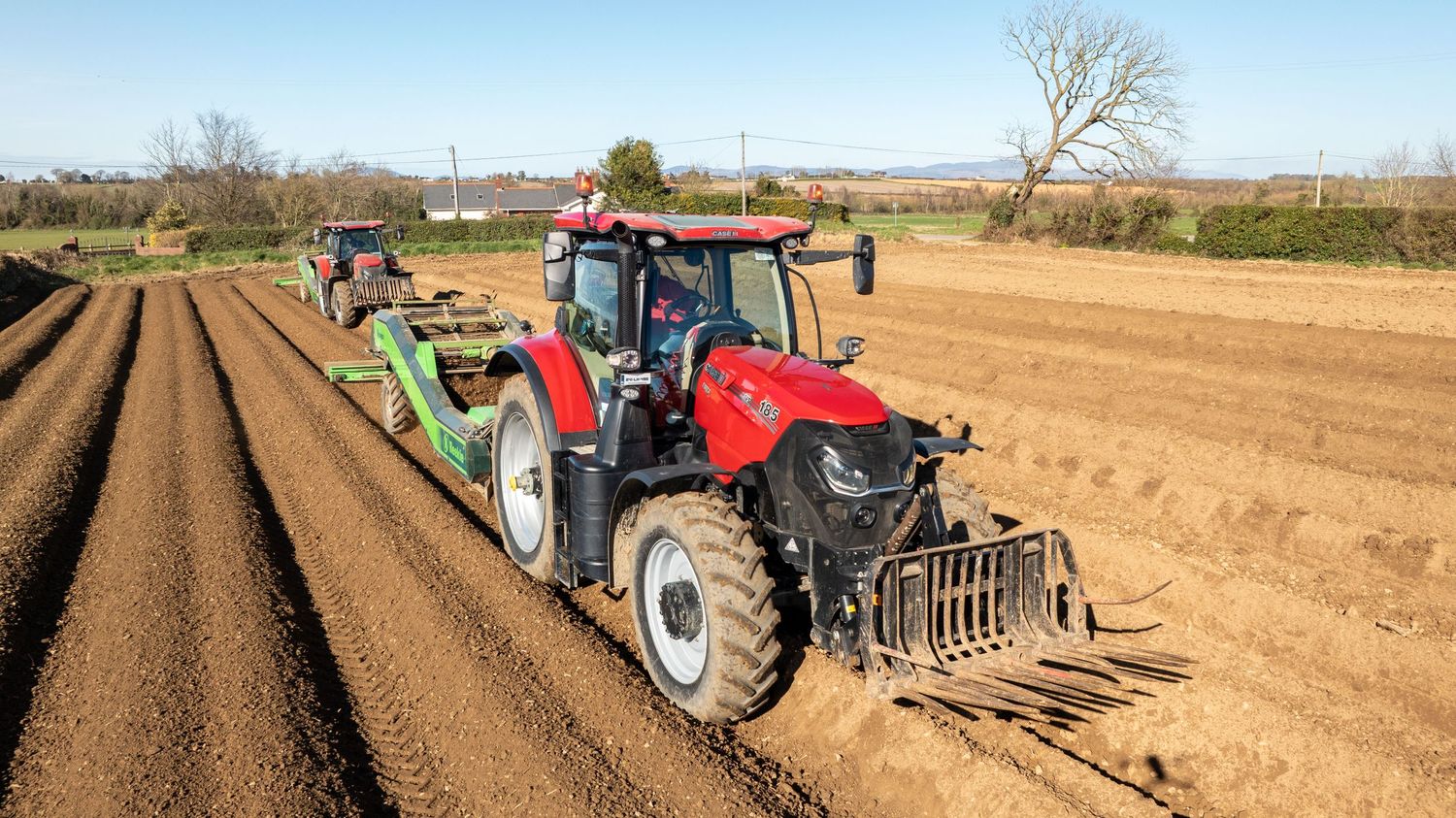Red tractor with farming equipment plows and prepares soil in agricultural field during spring planting season.