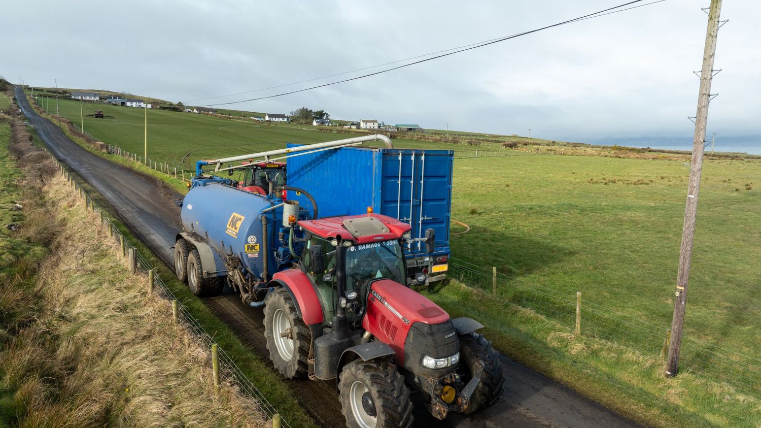 Red Case IH tractor pulling blue tanker trailers along rural road through green farmland under cloudy sky.