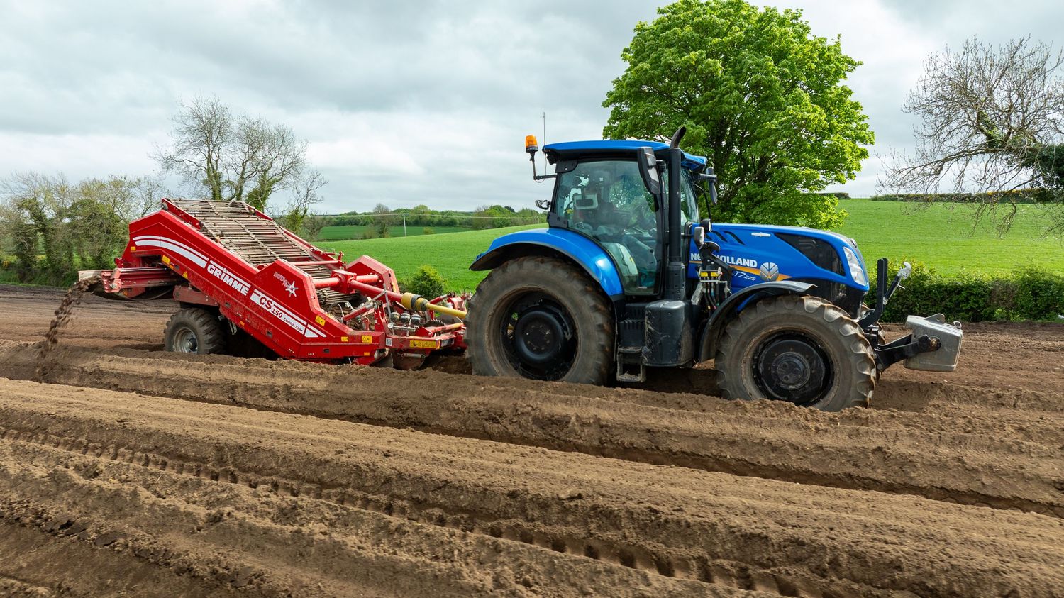 Blue agricultural tractor with red farming equipment working in plowed field under cloudy sky.