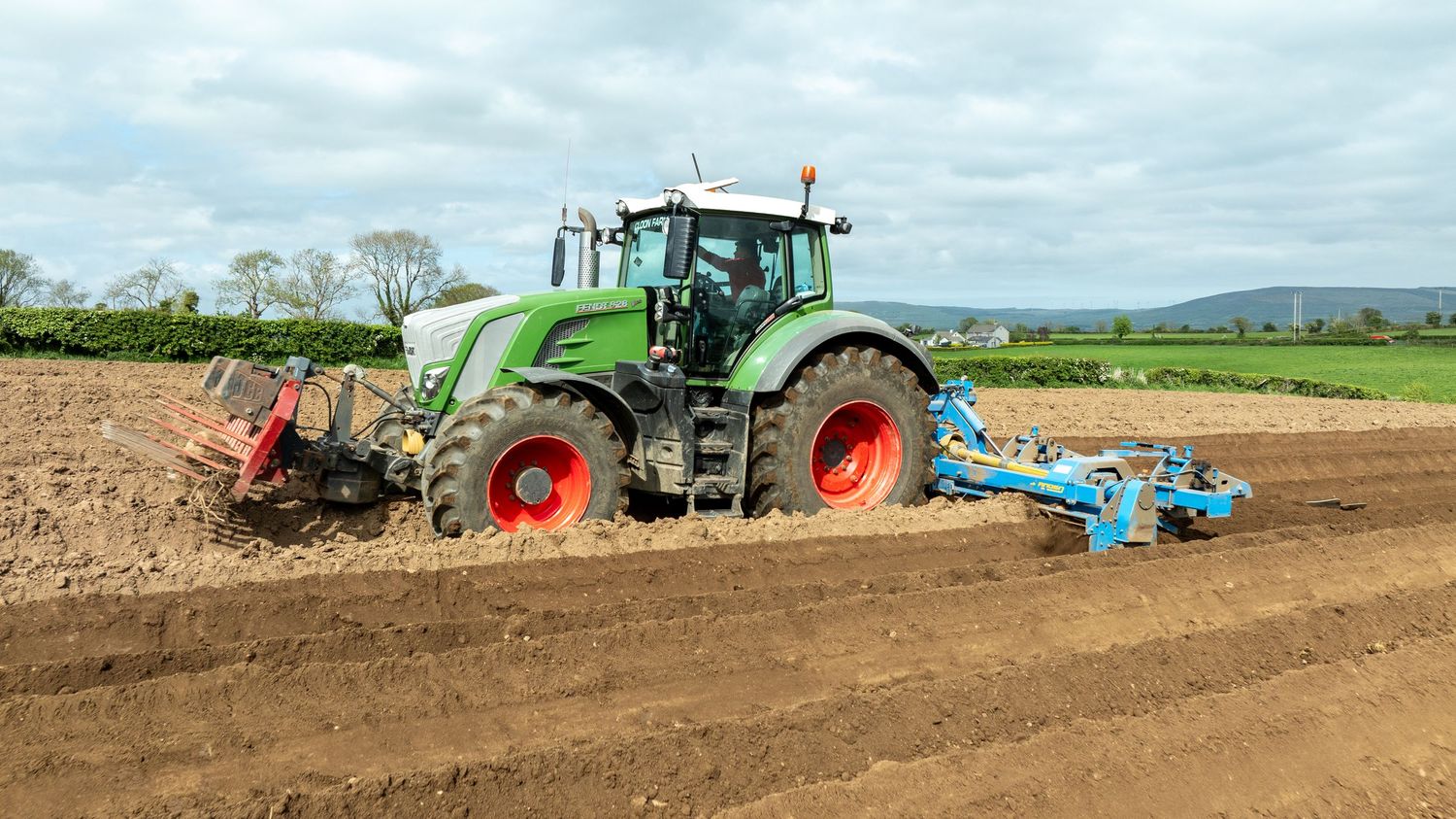 Green Fendt tractor pulling cultivator equipment across freshly tilled brown farmland on cloudy day.