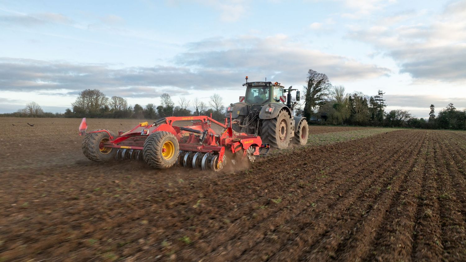 Tractor pulling red agricultural equipment across freshly plowed farmland under cloudy sky at dusk.