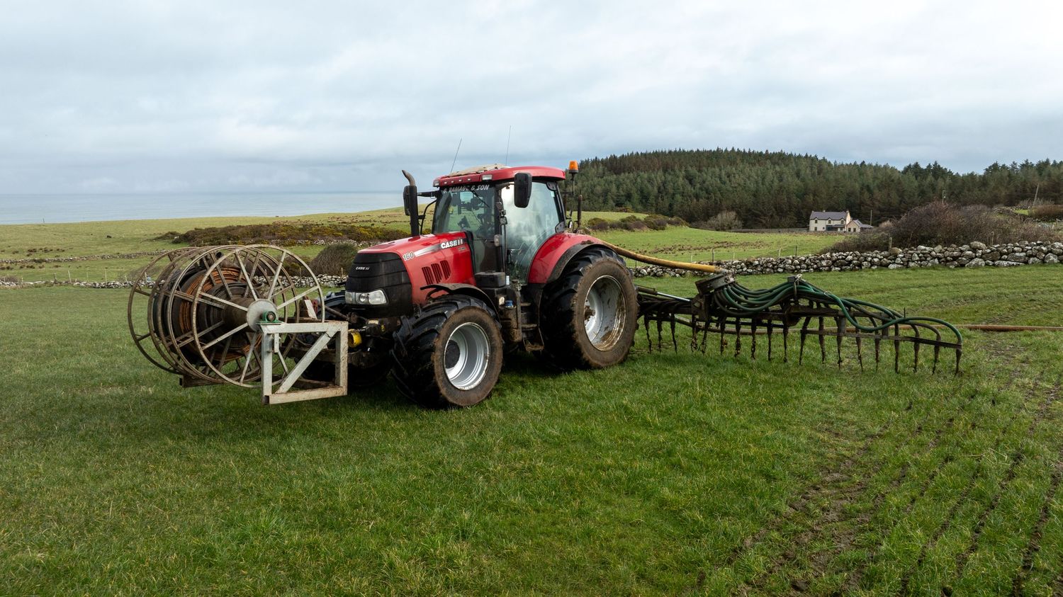 Red tractor pulling large metal farming equipment across green grassland with forested hills in the background.