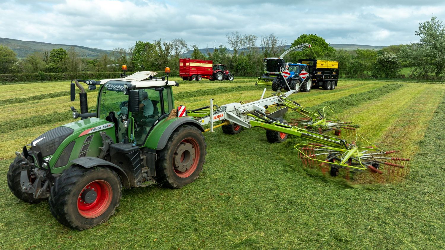 Agricultural machinery working in a green field with tractors and hay equipment harvesting grass on rolling farmland.