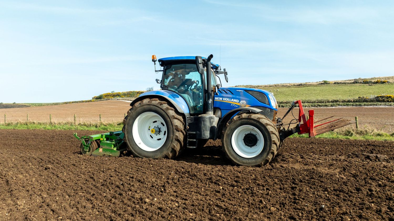 Blue New Holland tractor plowing farmland with agricultural equipment on a sunny day with clear blue sky.