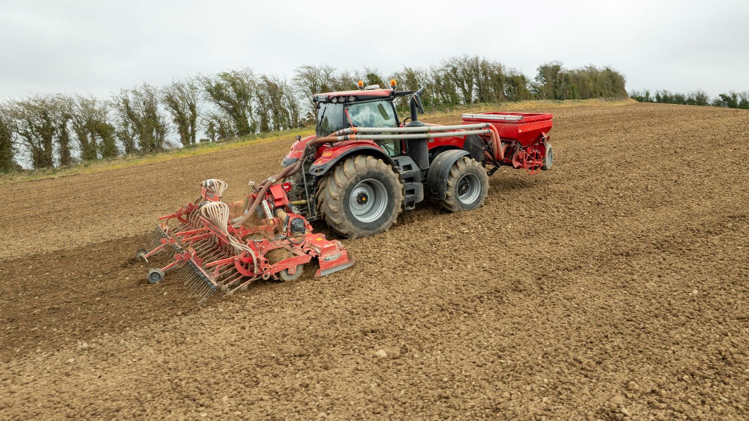 Red tractor with farming equipment tows seed and fertilizer spreaders across a freshly tilled brown field.