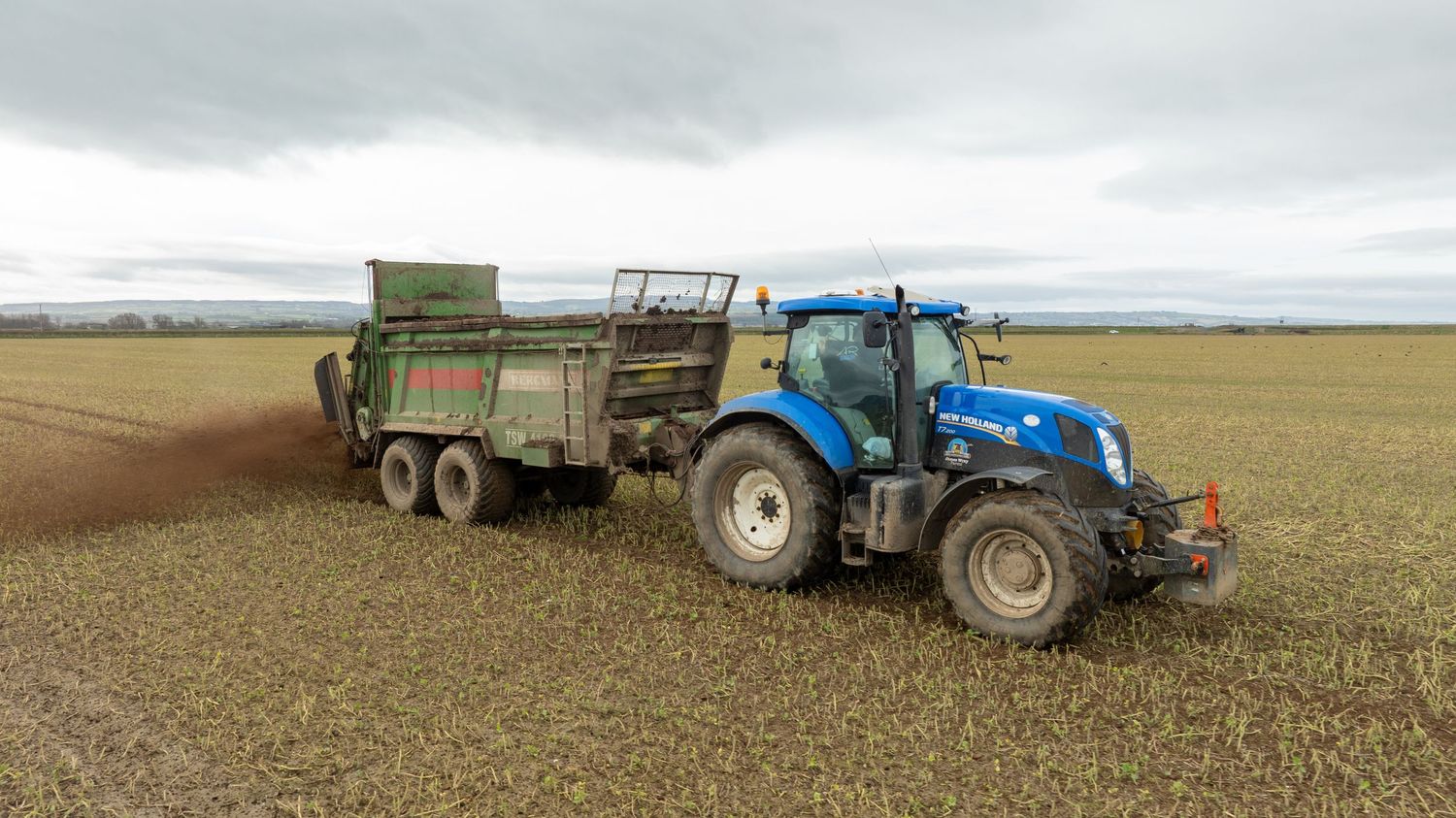 Blue New Holland tractor pulling a green trailer spreads manure across a harvested agricultural field under cloudy skies.