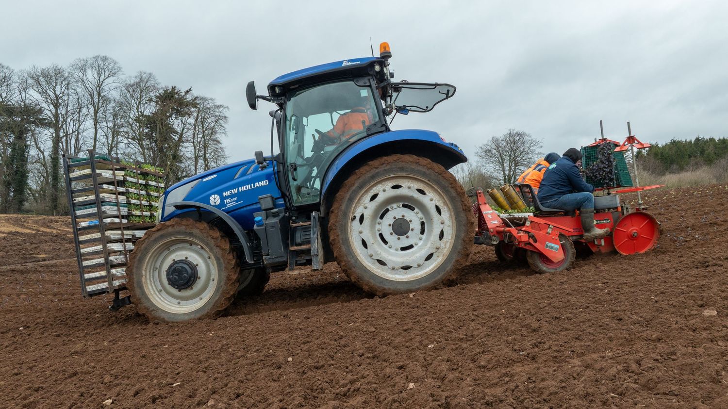 Blue tractor with attachments plowing dark soil in a rural farm field under cloudy skies.