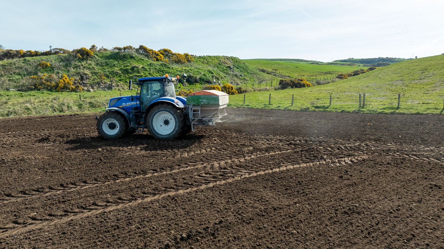 Blue tractor cultivating dark soil in rural farmland with green rolling hills in the background.