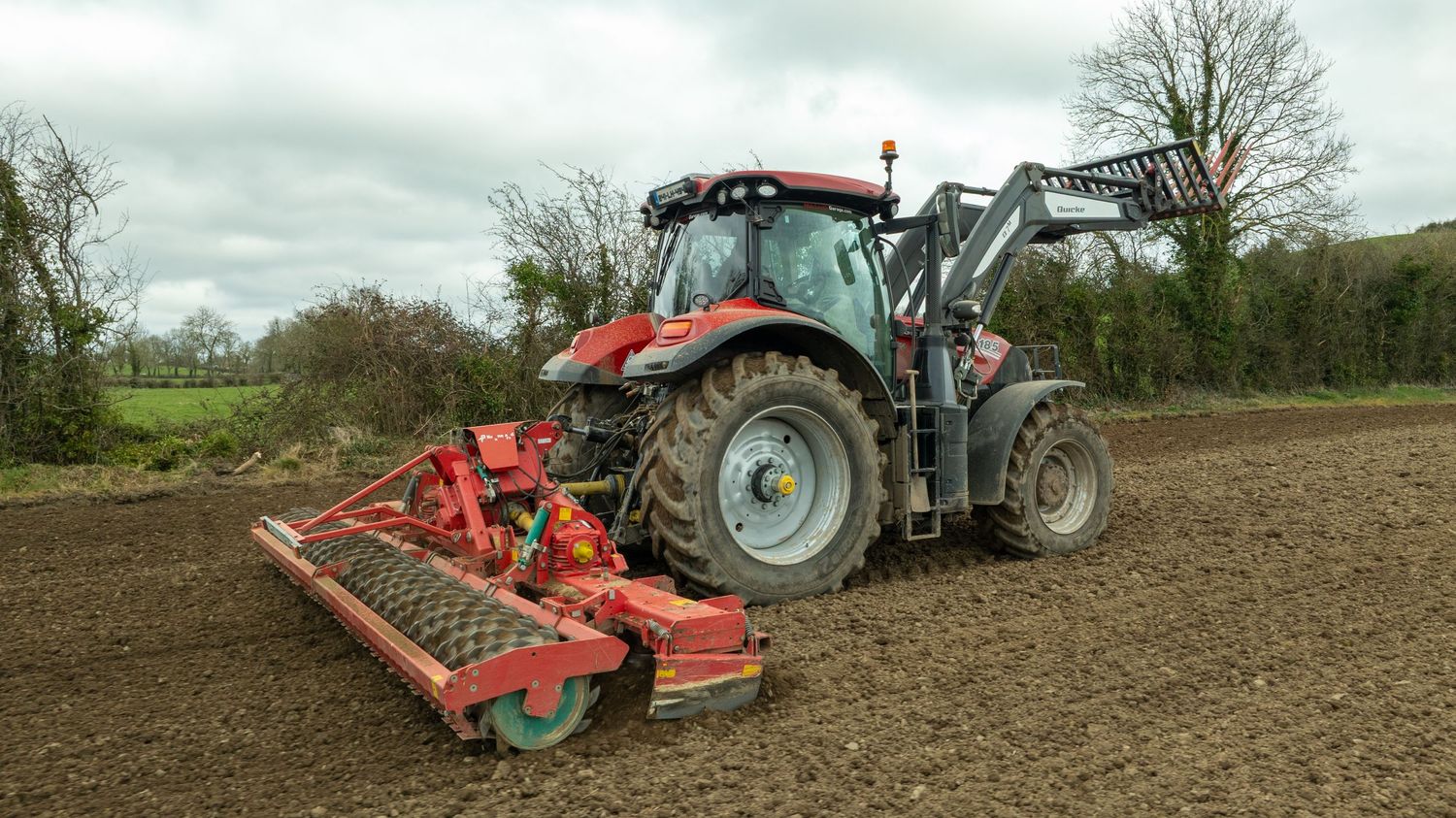 Red tractor with front loader and cultivator attachment working in a freshly plowed field under cloudy skies.