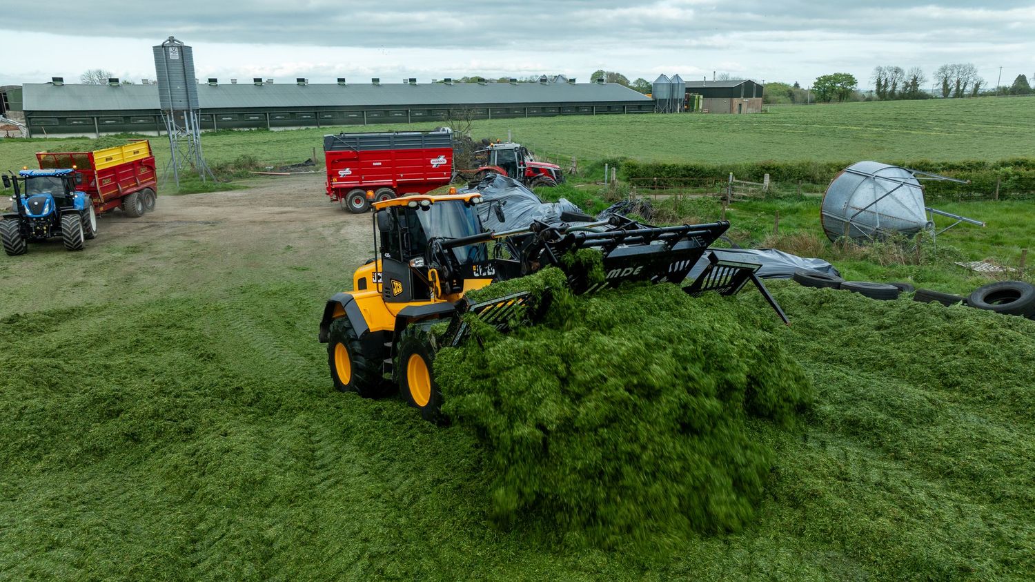 A yellow tractor parked on green grass with farm equipment and agricultural machinery in the background on a cloudy day.