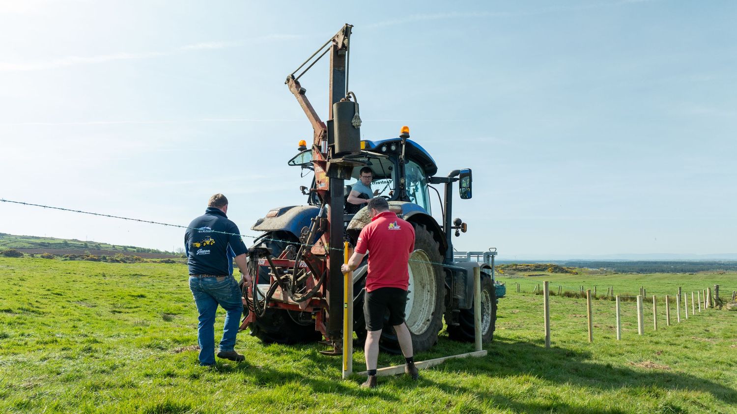 Farmers operate a post driver machine attached to a tractor on green pastureland with fencing in the background.