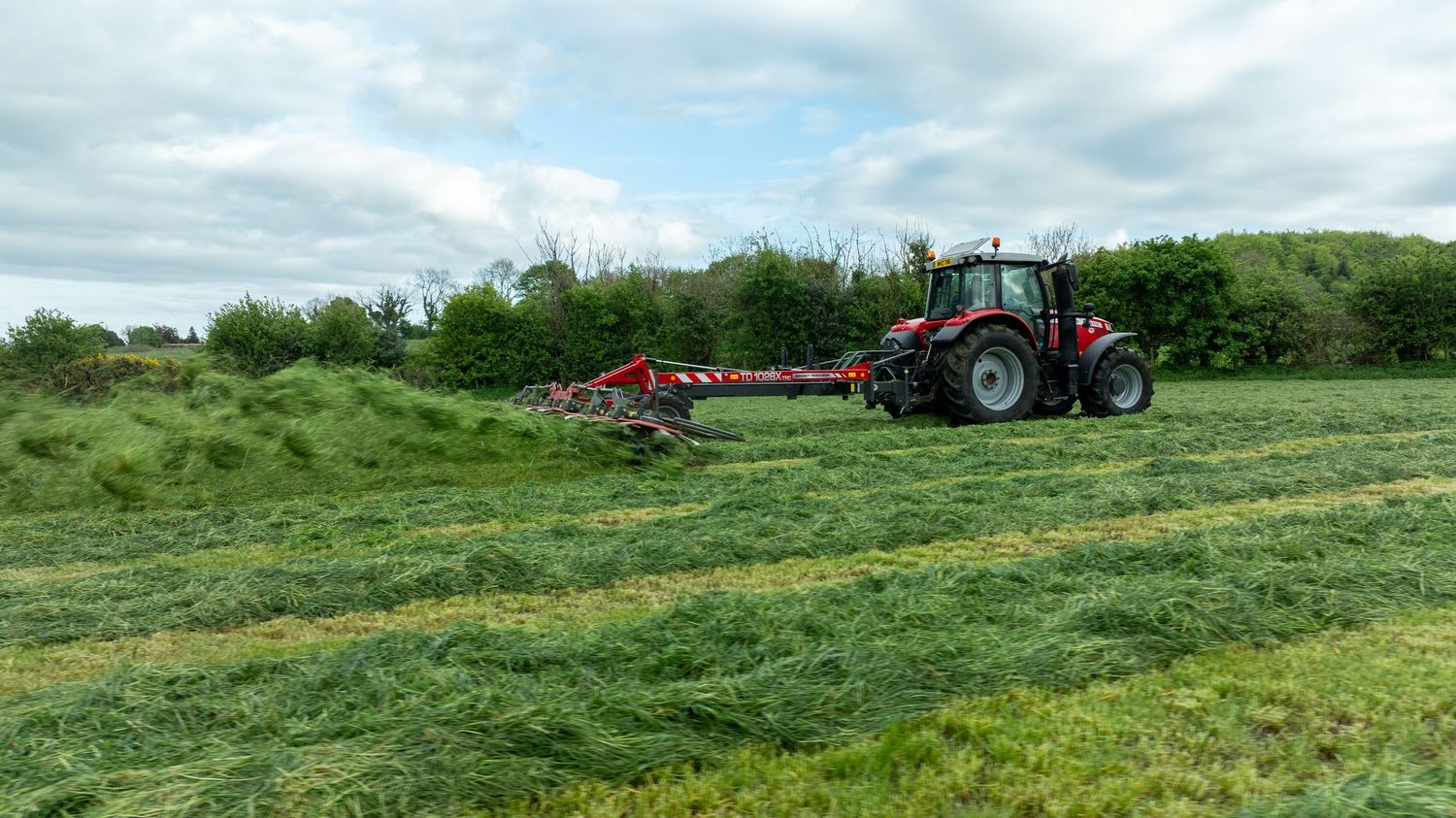 Red tractor pulling hay tedding equipment across a lush green field on a cloudy day.