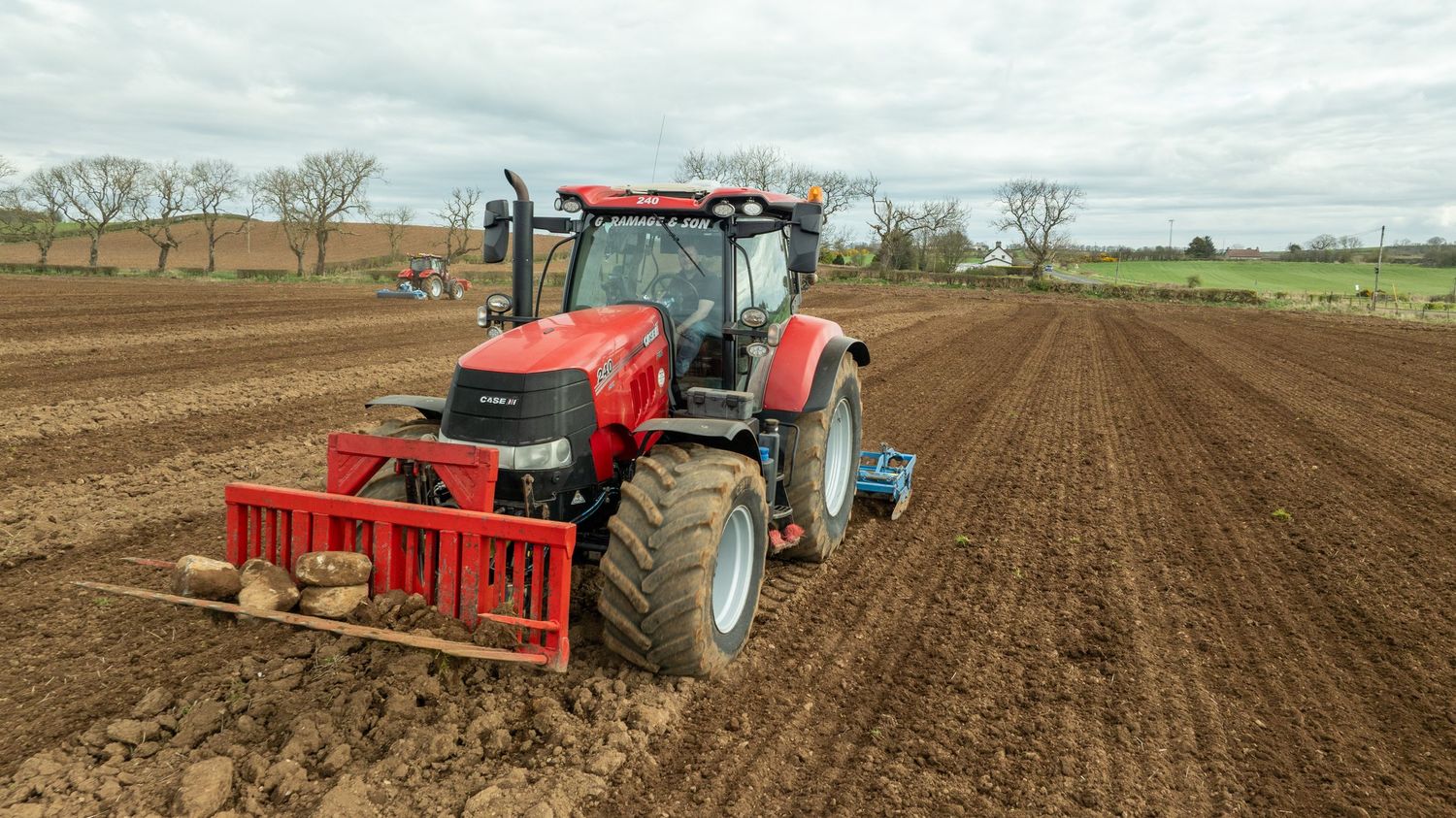 Red tractor with cultivator attachment plowing rows in a brown agricultural field under cloudy skies.