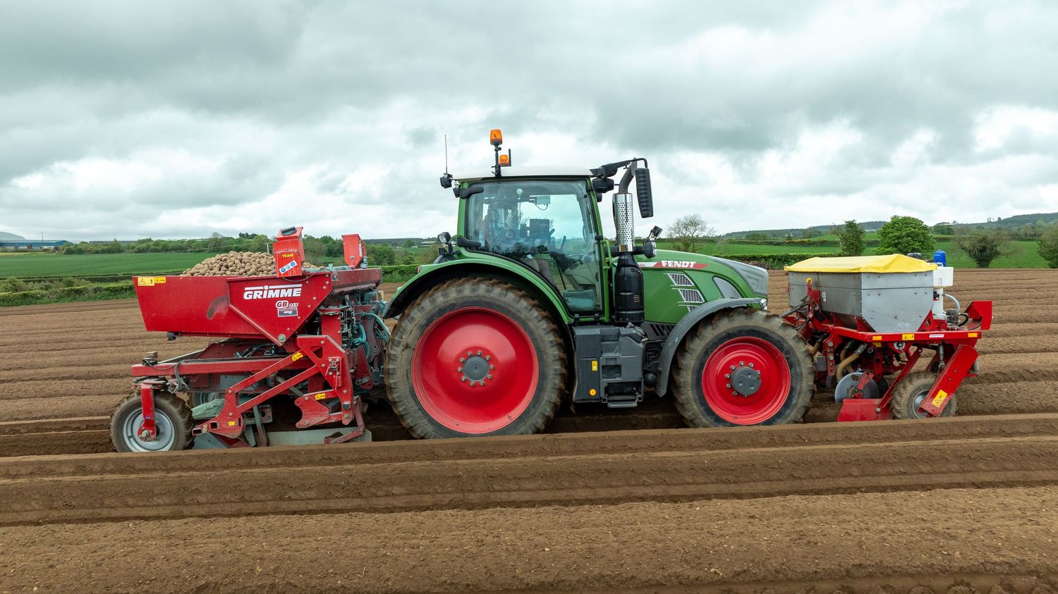 Large green tractor pulling red agricultural planting equipment across freshly tilled soil under cloudy skies.