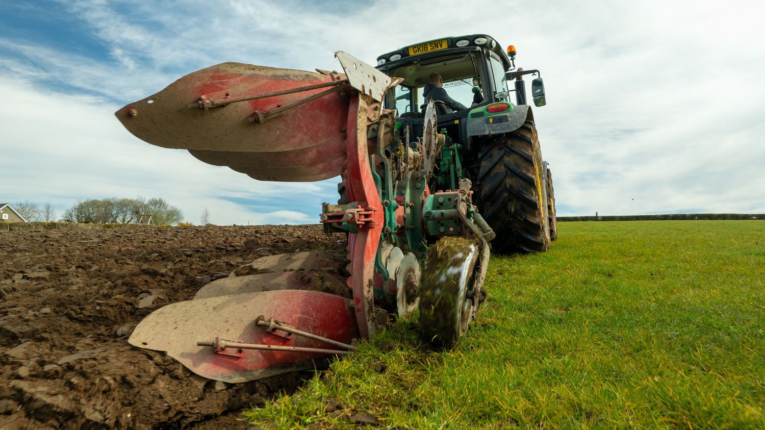 A tractor with plow attachment works at the edge of a field, creating a line between plowed soil and green grass.