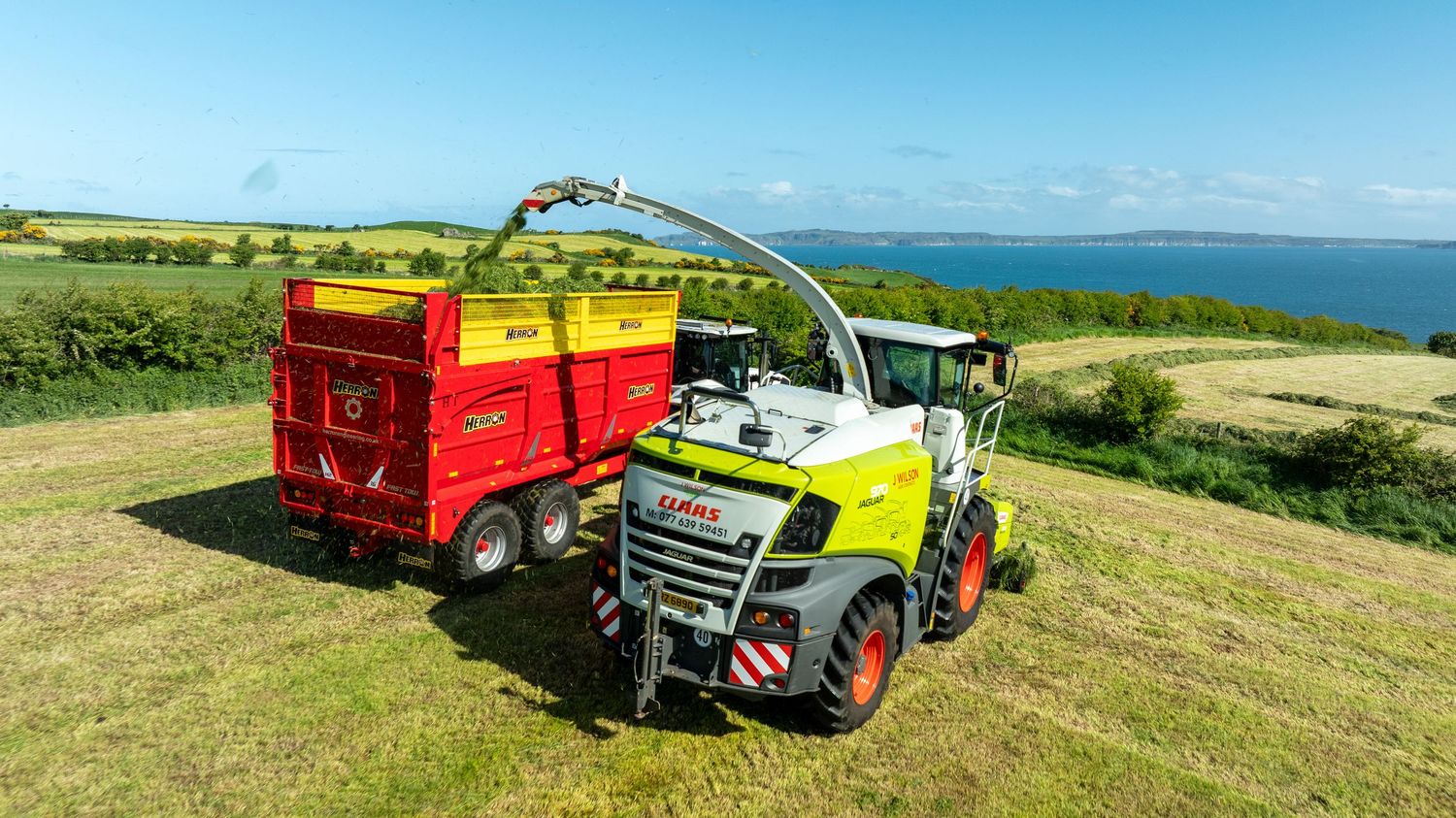 Agricultural machinery harvesting green grass silage along coastal fields with bright blue ocean view in background.
