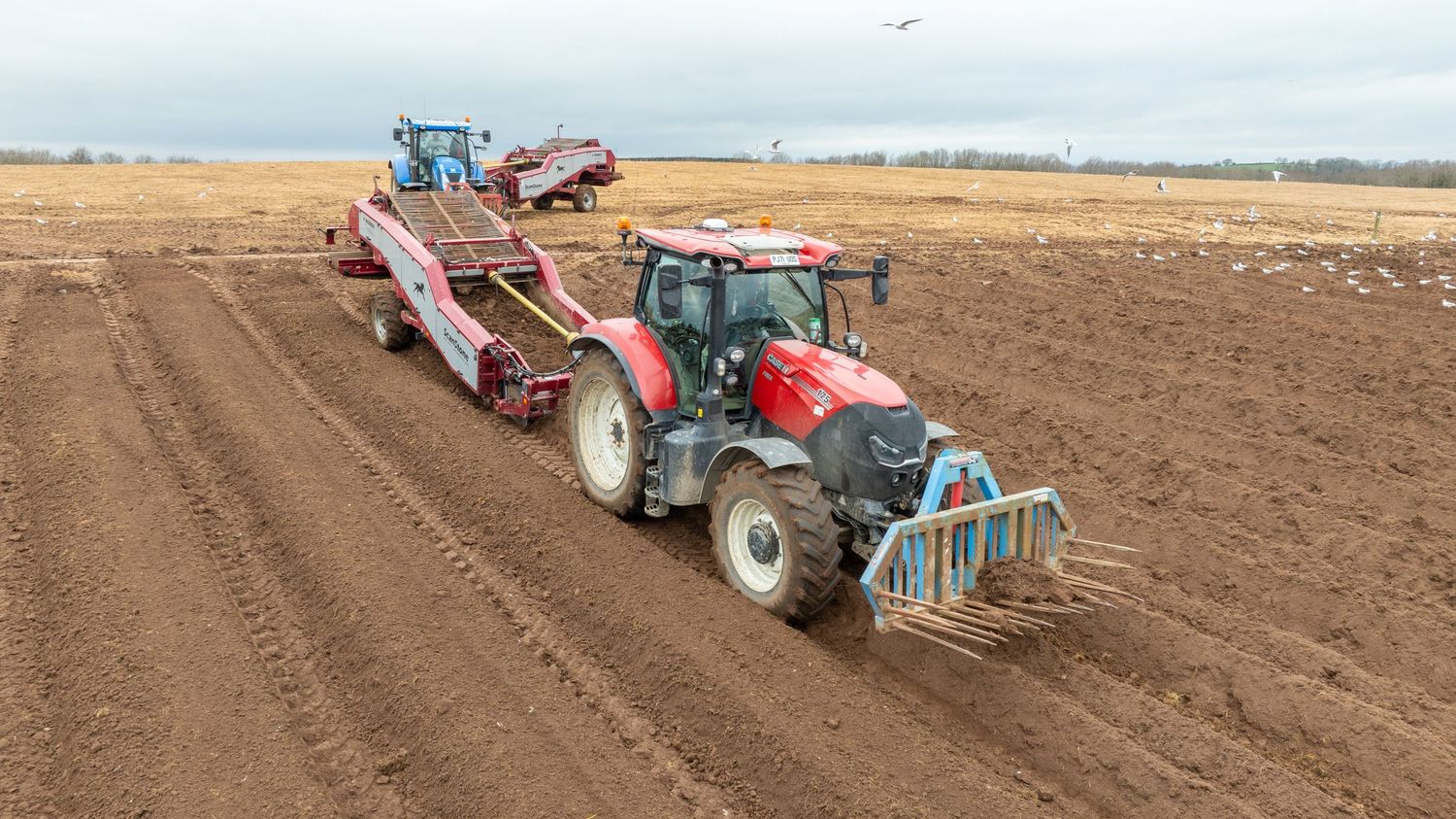 Red tractor pulling agricultural equipment plows brown soil in a farm field under cloudy skies.