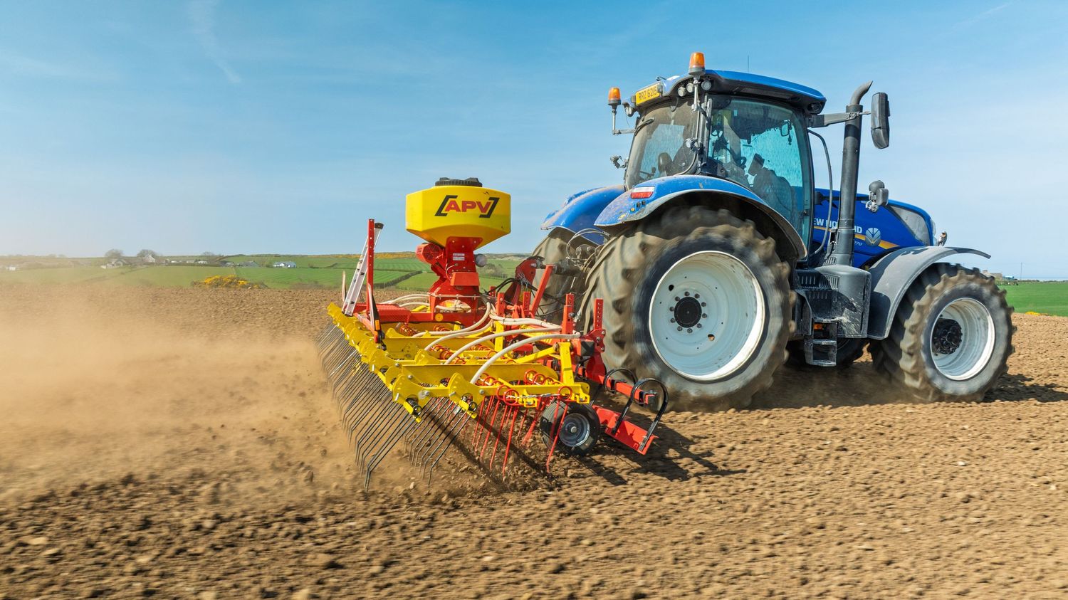 Blue tractor with yellow seeding equipment plowing and planting crops in dry dusty agricultural field on sunny day.