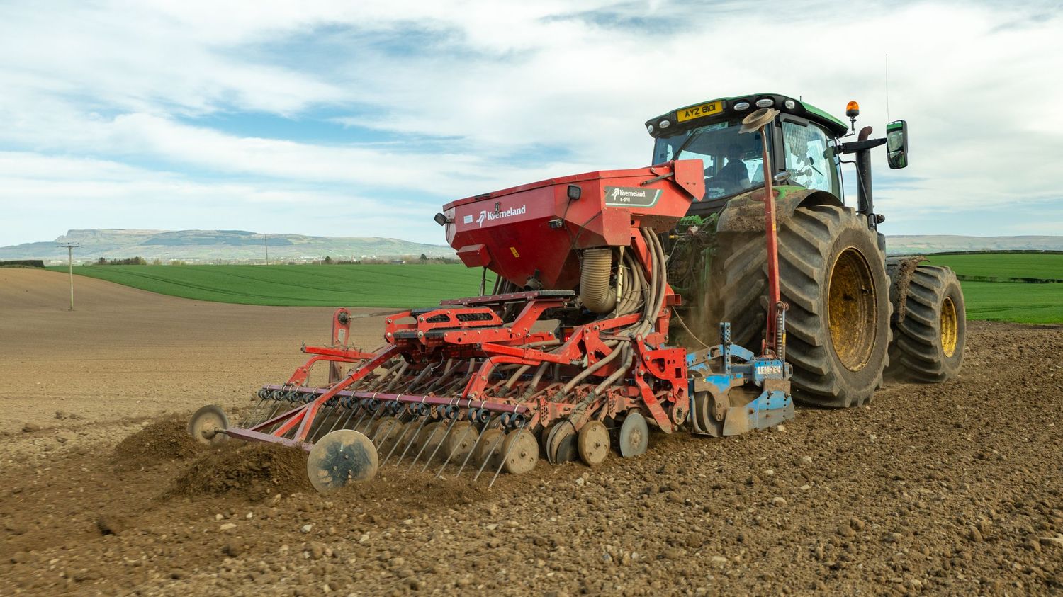 Orange tractor with modern seed drill equipment cultivating and seeding soil in agricultural field on cloudy day.