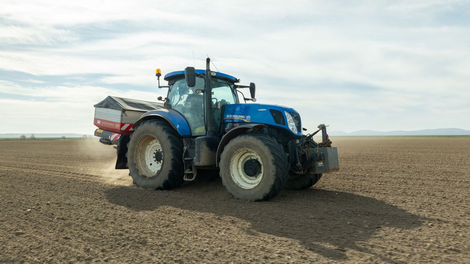 A blue New Holland tractor with fertilizer spreader working in a plowed agricultural field under cloudy skies.
