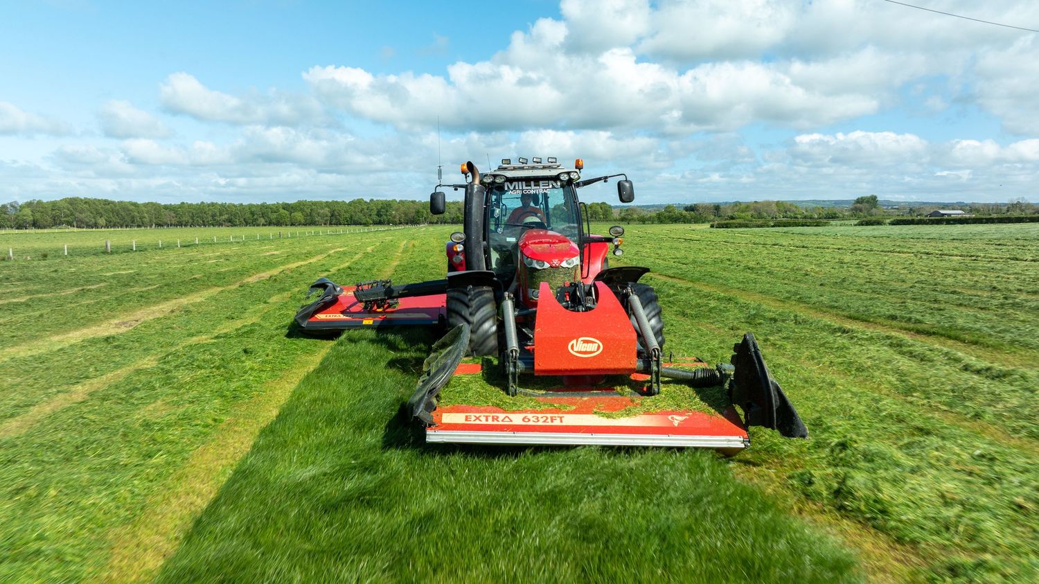 Two red tractors with mowing attachments cutting grass in a lush green field under a bright cloudy sky.