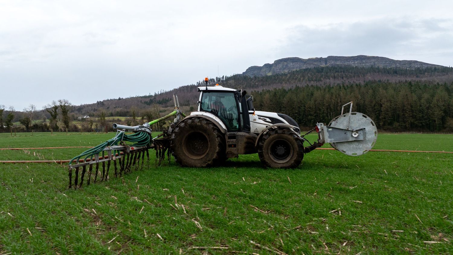 A green tractor with fertilizer equipment operates on a lush field with forested mountains in the background.