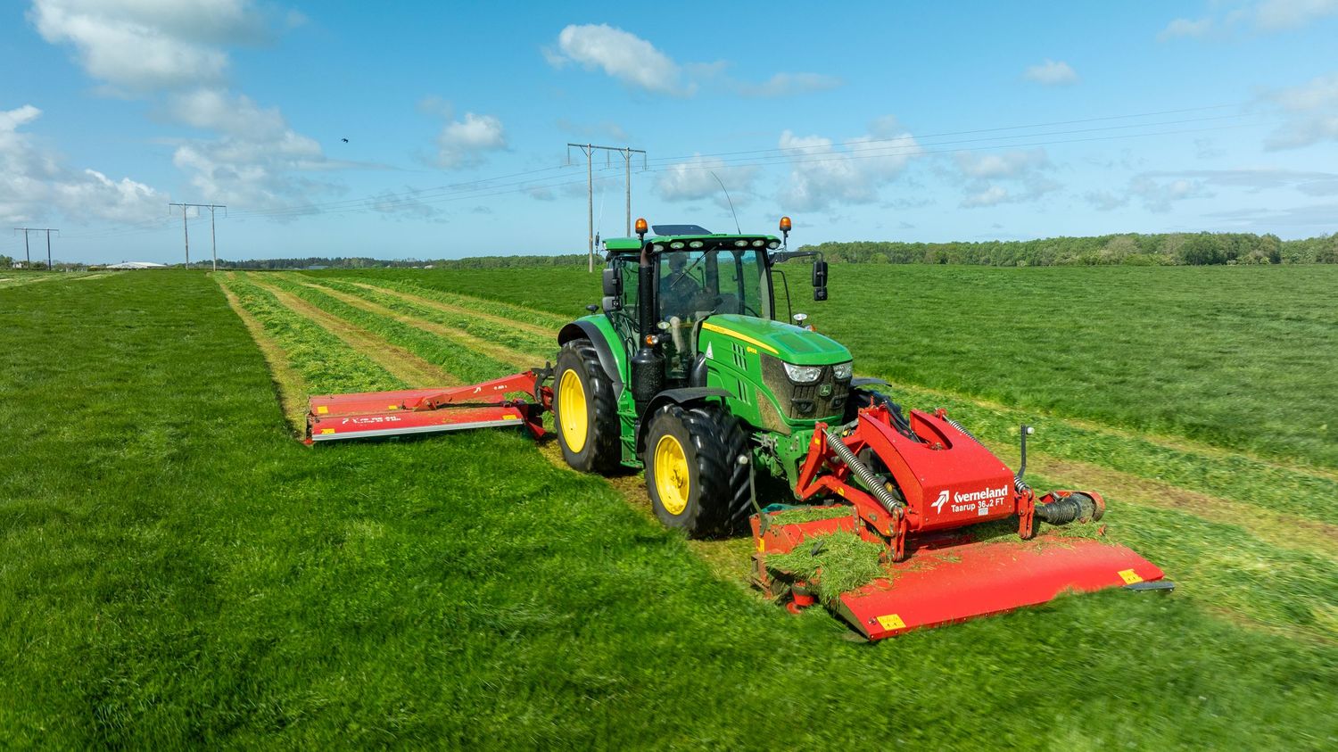 Green John Deere tractor mowing large grass field with two red mower attachments under blue sky with white clouds.
