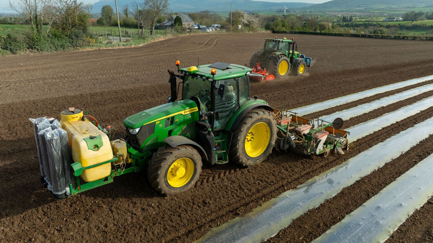 Two green John Deere tractors plowing and preparing agricultural field with plastic mulch laying equipment.