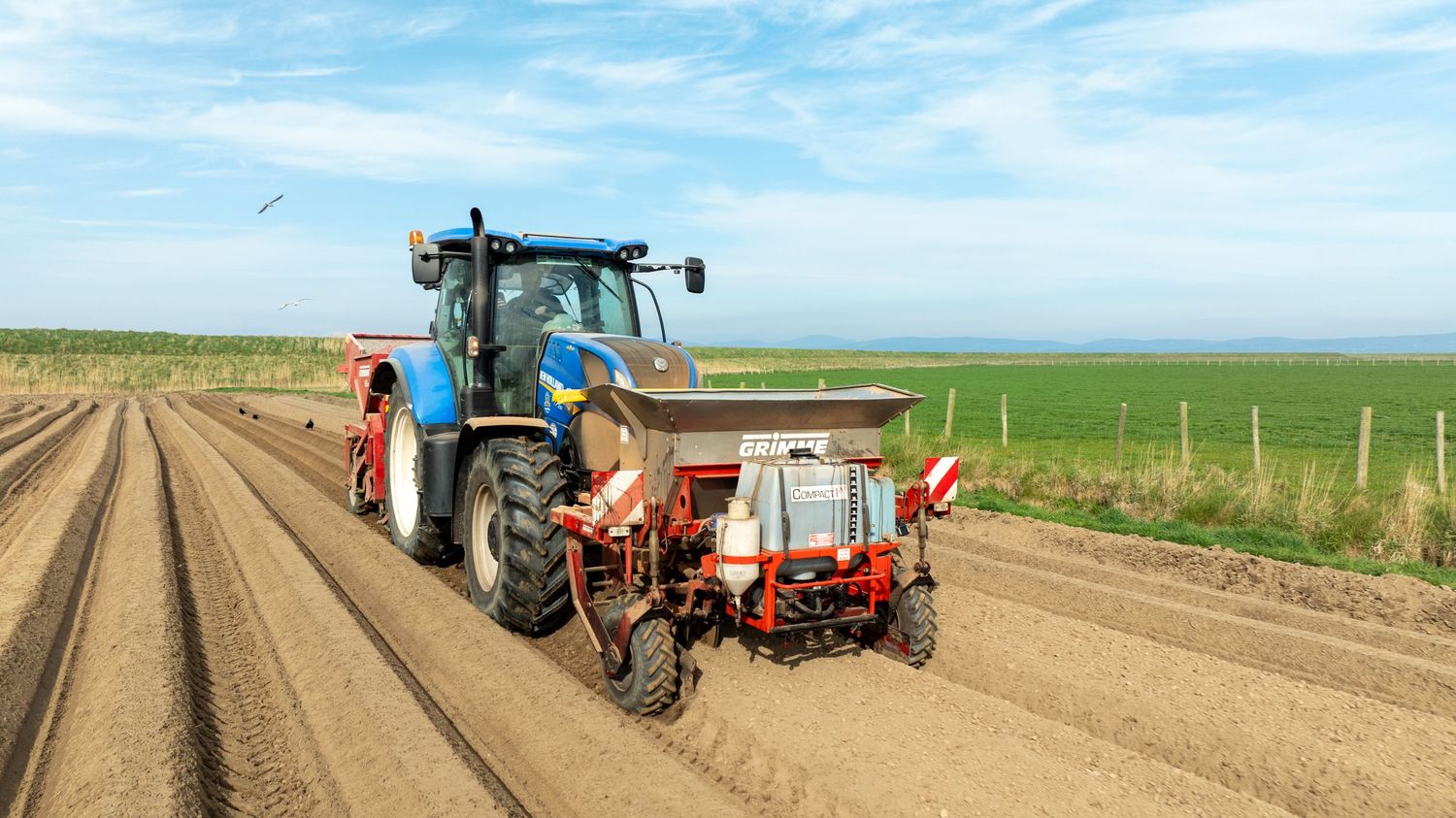 Blue tractor pulling farm equipment across plowed agricultural field under blue sky with farmland in background.
