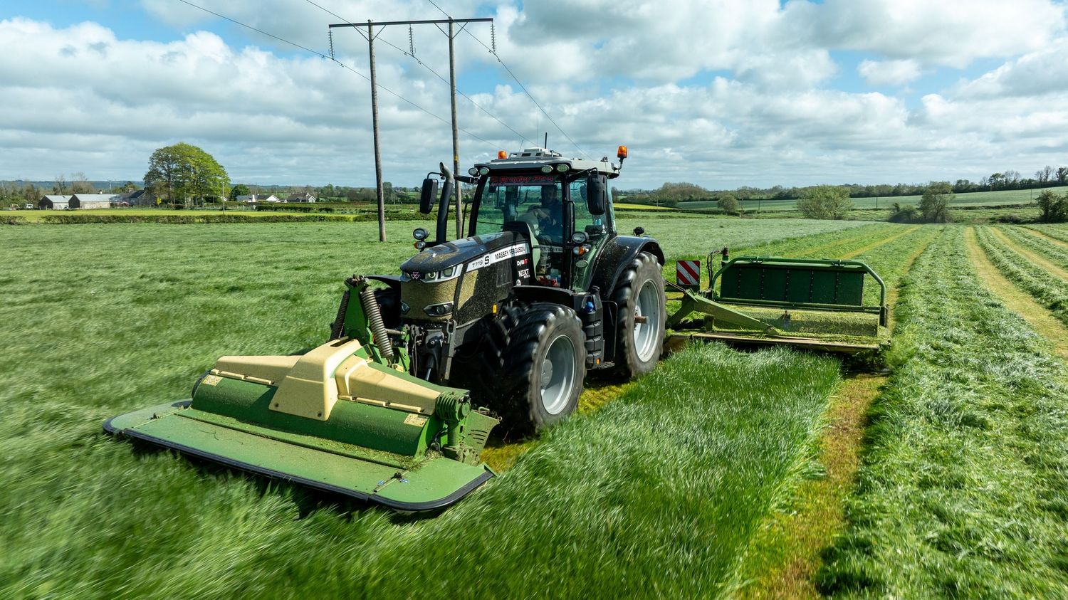 Modern green tractor mowing grass in a lush field with multiple mower attachments under cloudy summer sky.