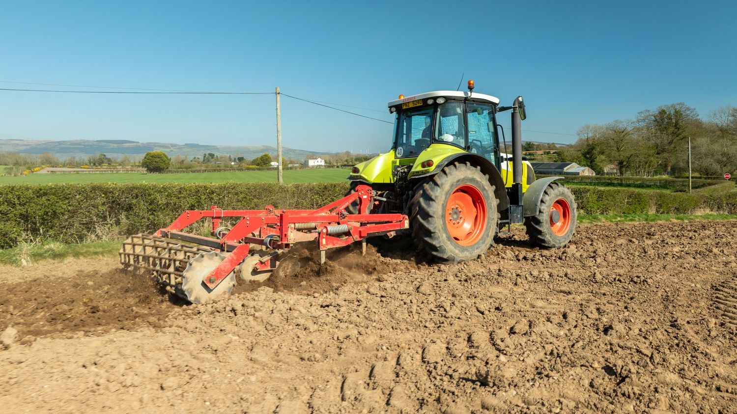 Green tractor pulling red cultivator equipment through plowed field on sunny day with blue sky in rural countryside.