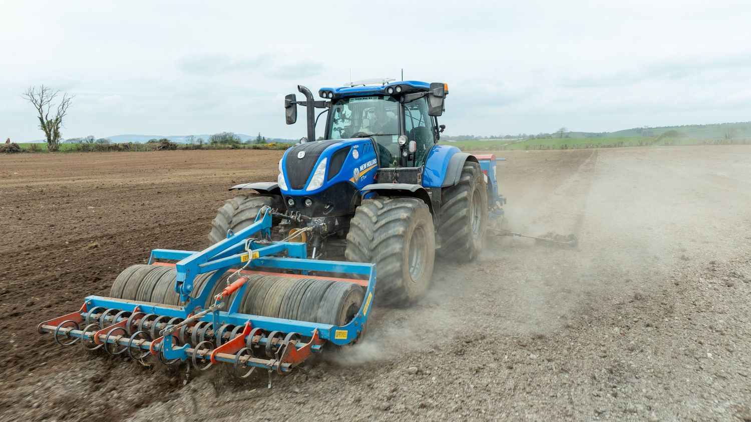 A blue New Holland tractor pulling a tillage implement works in a dusty field preparing soil for planting.