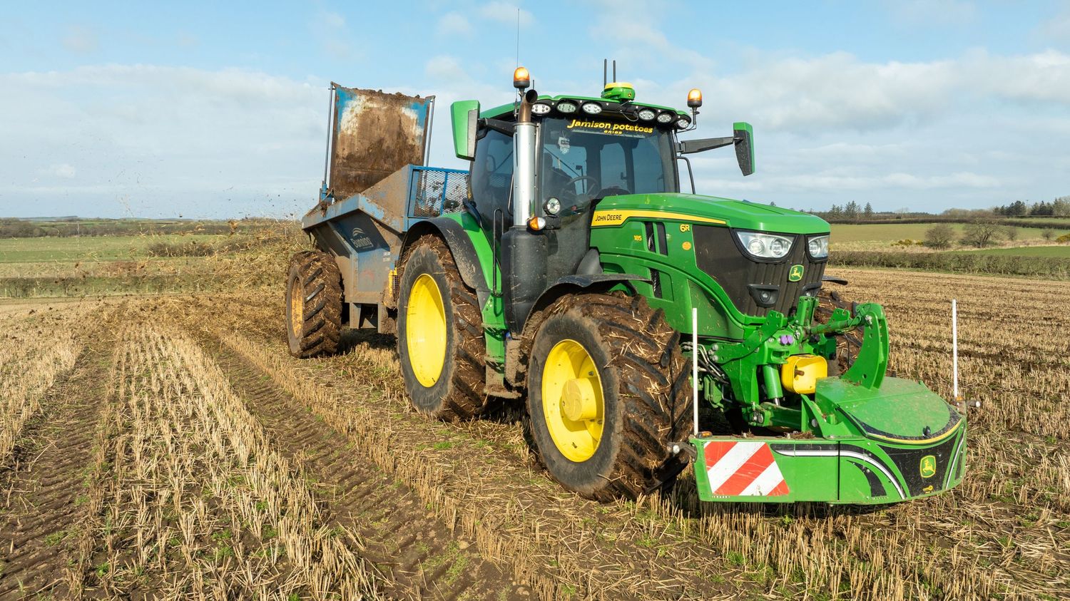 A green John Deere tractor pulling farm equipment across a harvested agricultural field on a sunny day.