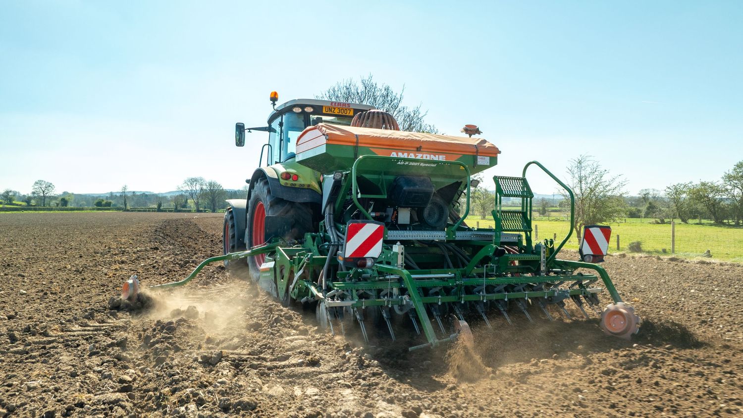 Large green farm machinery planting seeds in freshly tilled soil with dust trailing behind on a sunny spring day.