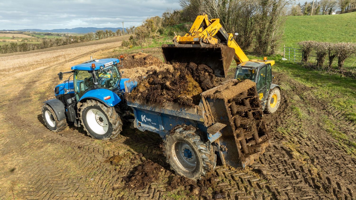Blue tractor and yellow excavator working together to move soil on a dirt road with rolling hills in background.