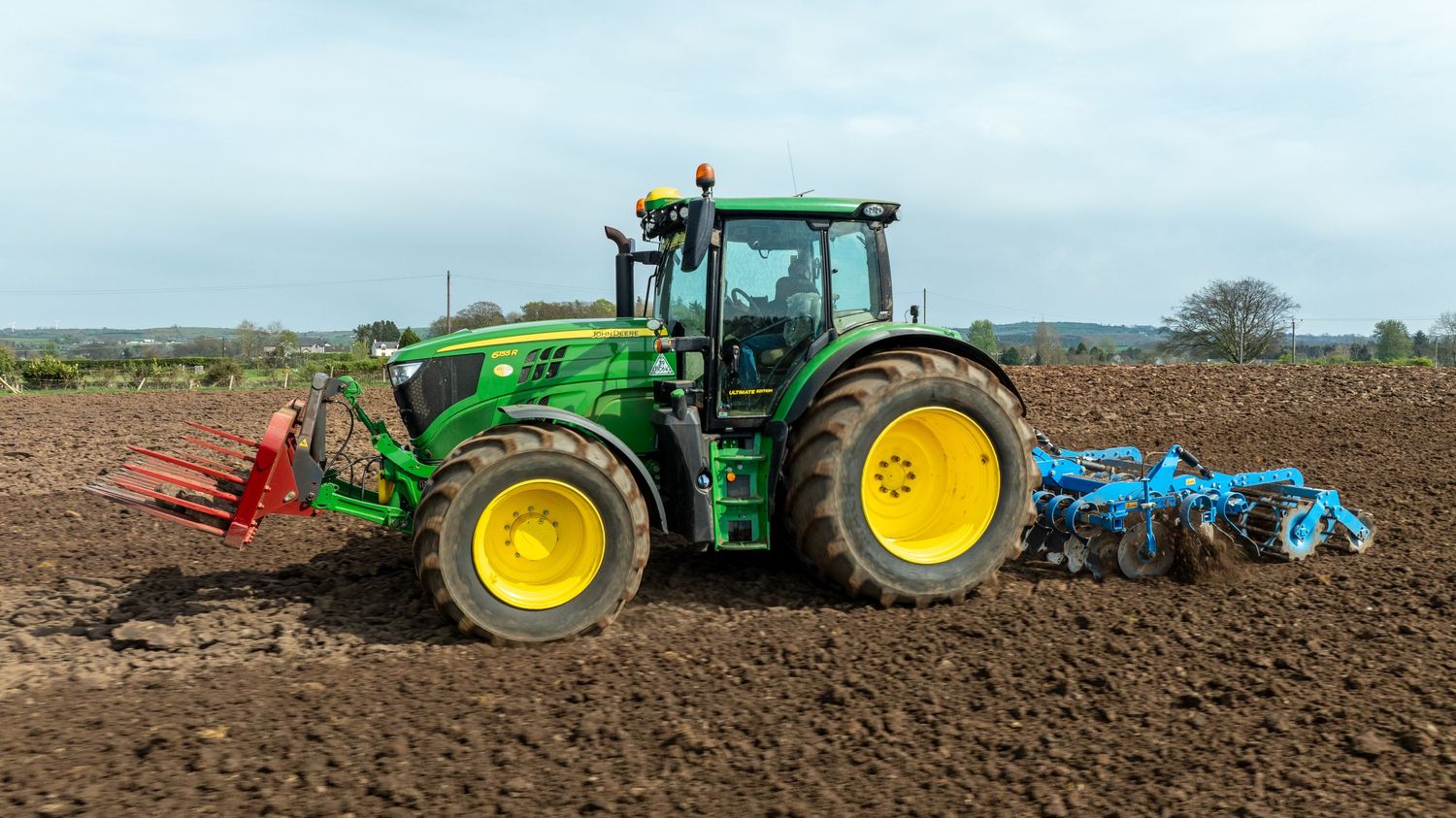 A green John Deere tractor with blue and red implements plows through dark soil in a rural farm field.